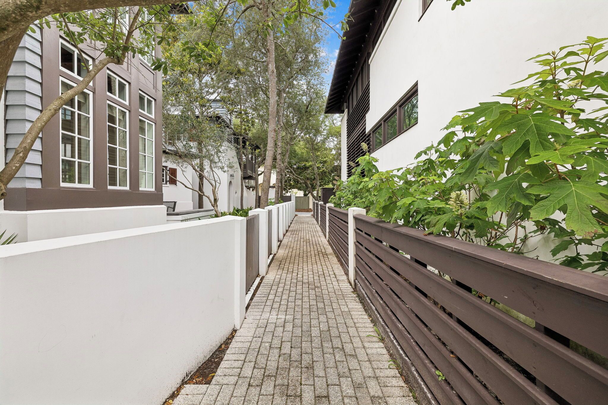 A narrow pathway flanked by white walls and a wooden fence, leading to a building with large windows and a dark roof.