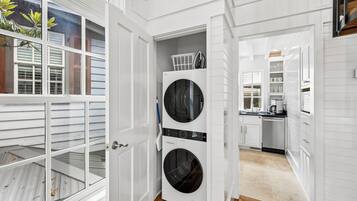 A laundry room with a front-loading washer and dryer, a small laundry basket on top, and a view into the adjacent kitchen area.