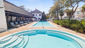 En swimmingpool med solstolar och en pergola, omgiven av träd och byggnader.