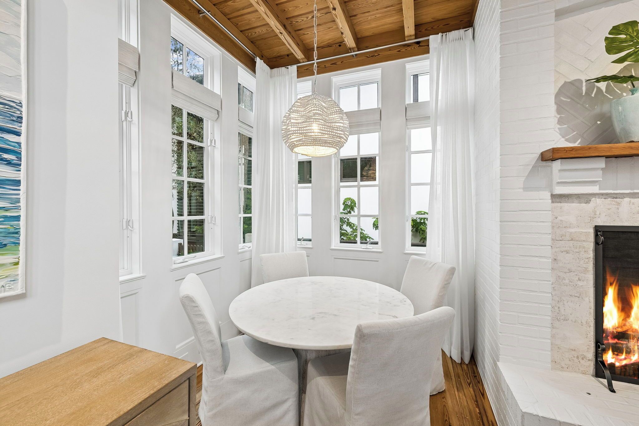 A room with a round table and chairs, large windows, a fireplace, and a hanging light fixture.