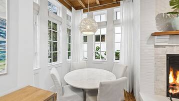 A room with a round table and chairs, large windows, a fireplace, and a hanging light fixture.