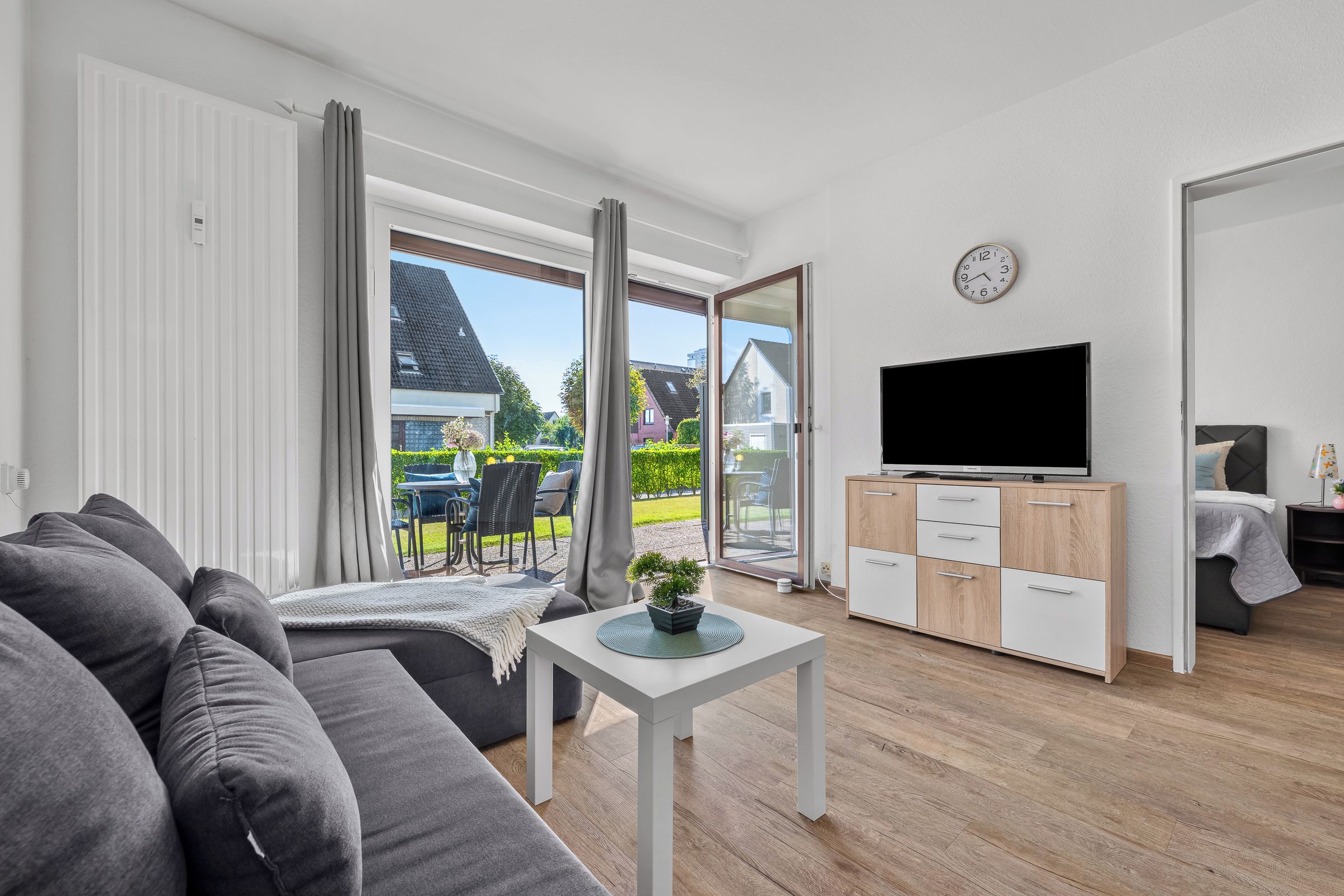 Un salon moderne avec un canapé gris, une petite table blanche ornée d’une plante, un téléviseur posé sur un meuble en bois et une porte-fenêtre coulissante donnant sur une terrasse extérieure.