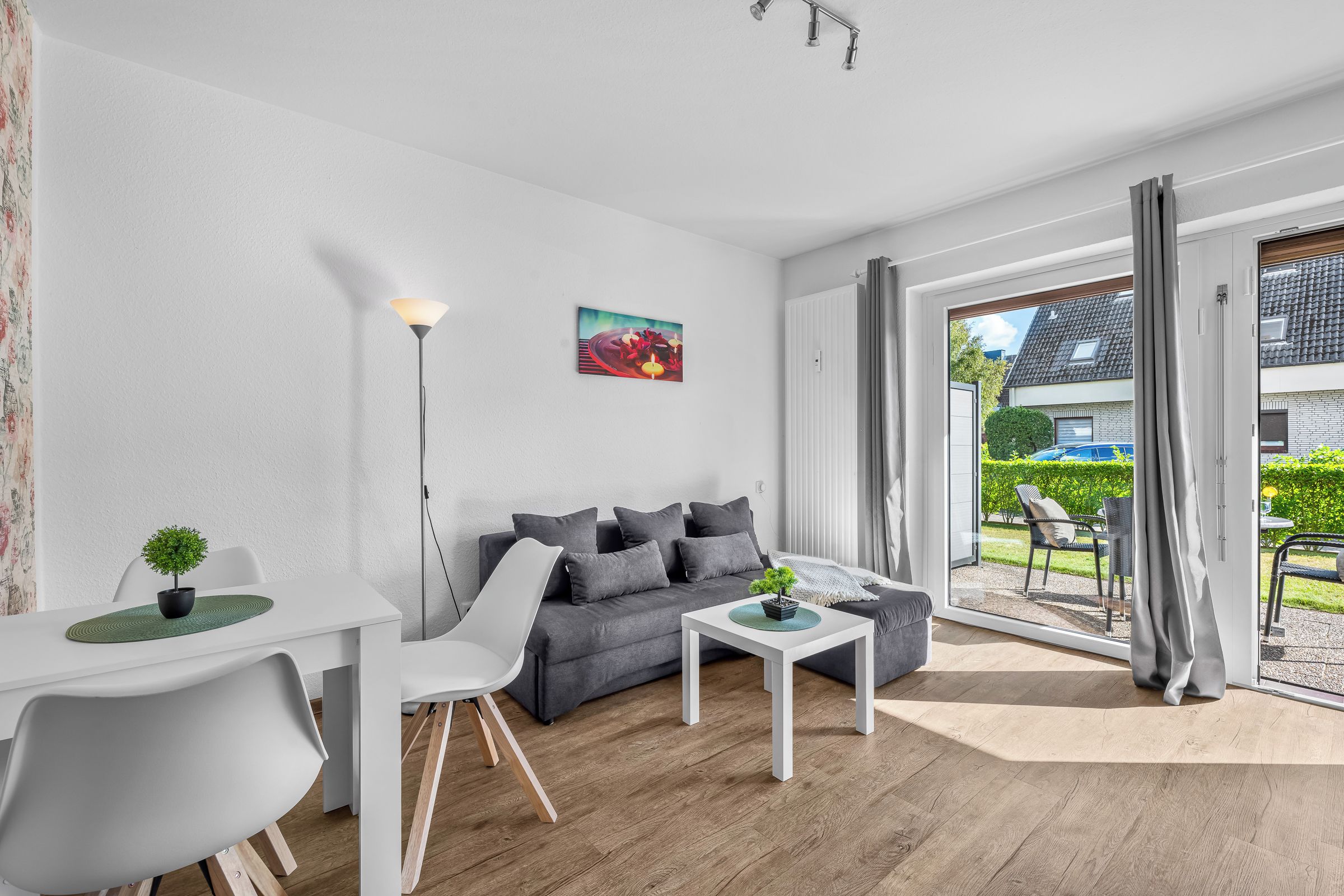 Una sala de estar moderna con un sofá gris, una mesa de centro blanca, una lámpara de pie y una zona de comedor con una mesa y sillas blancas.