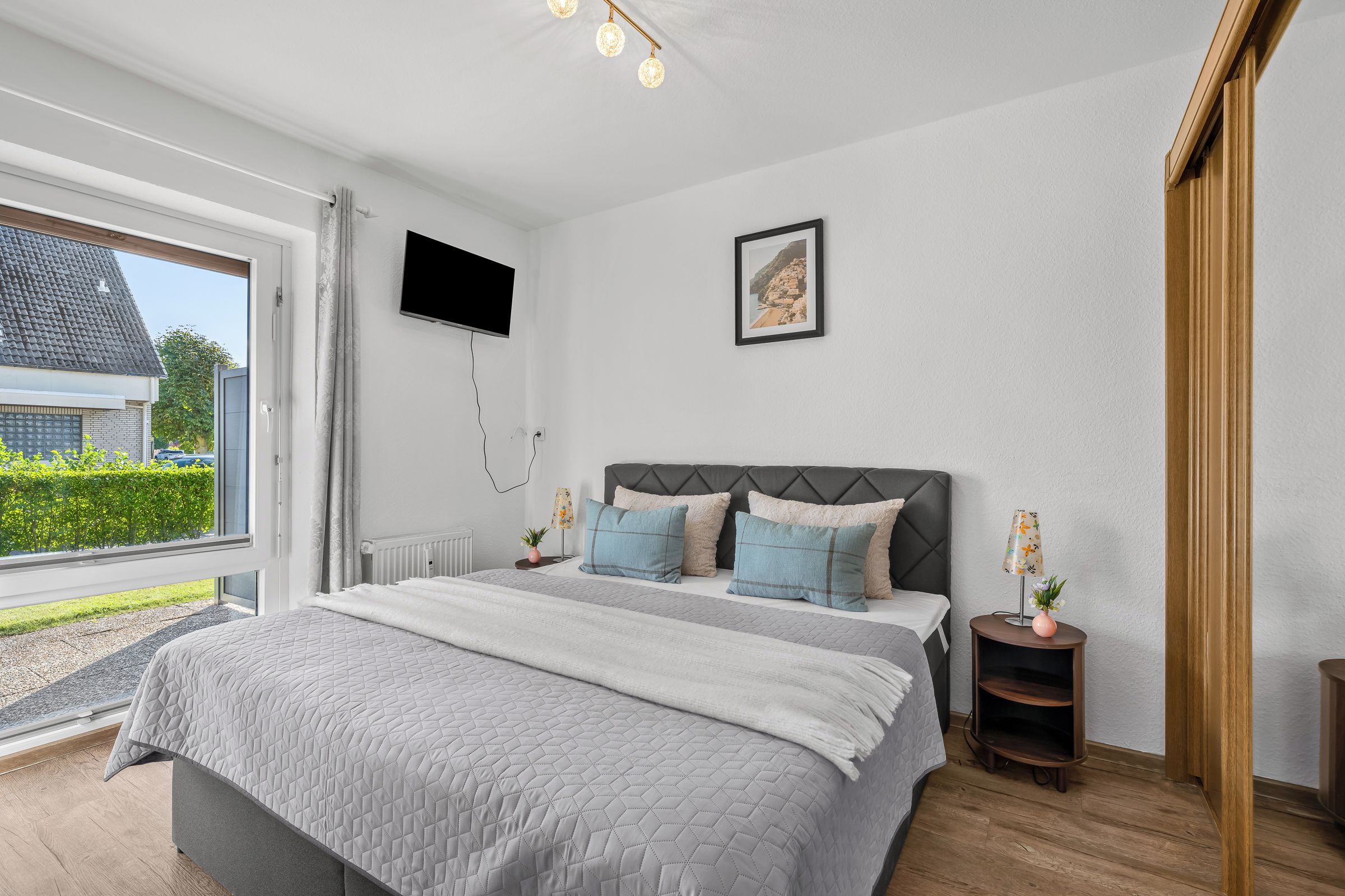Ein Schlafzimmer mit Bett, einem an der Wand montierten Fernseher, einem gerahmten Bild und einem Fenster mit Blick auf ein Haus und Grünflächen.
