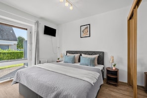 Ein Schlafzimmer mit Bett, einem an der Wand montierten Fernseher, einem gerahmten Bild und einem Fenster mit Blick auf ein Haus und Grünflächen.