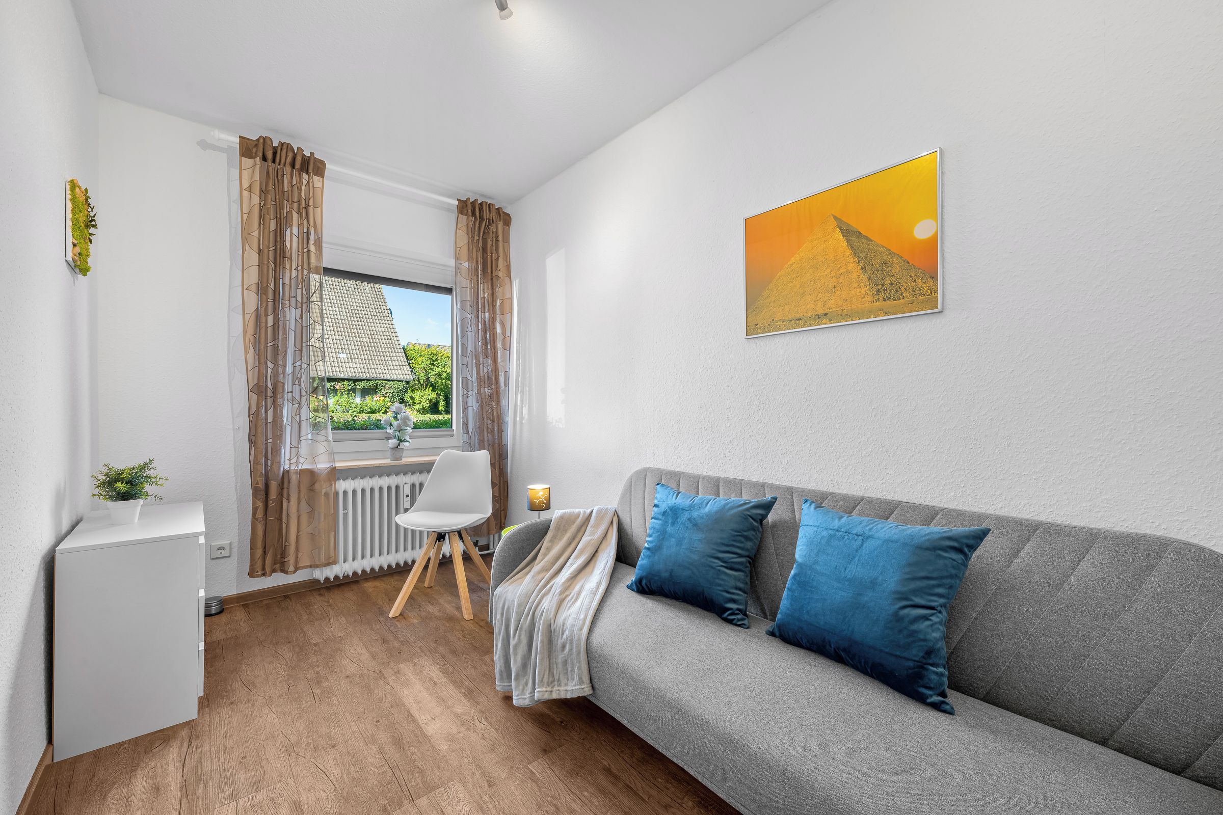 A modern living room with a grey sofa, blue cushions, a small white desk, and a window with curtains.