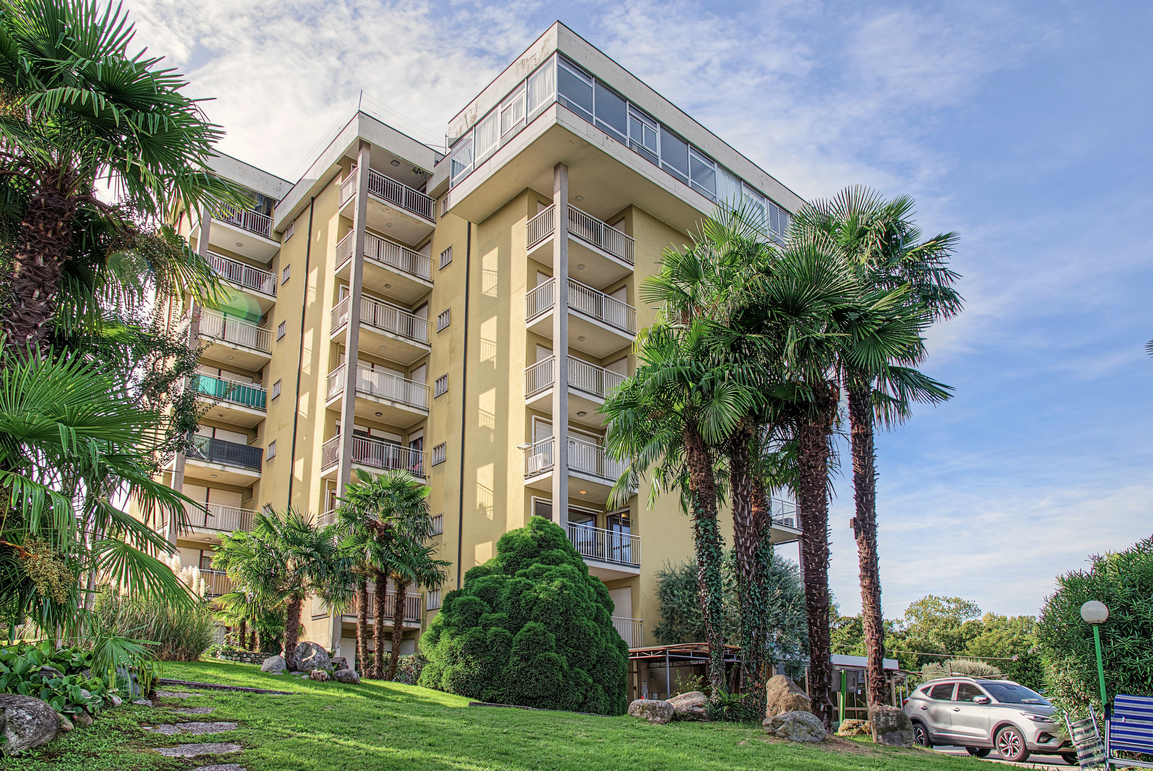 A multi-story residential building with balconies, surrounded by palm trees and a well-maintained lawn.