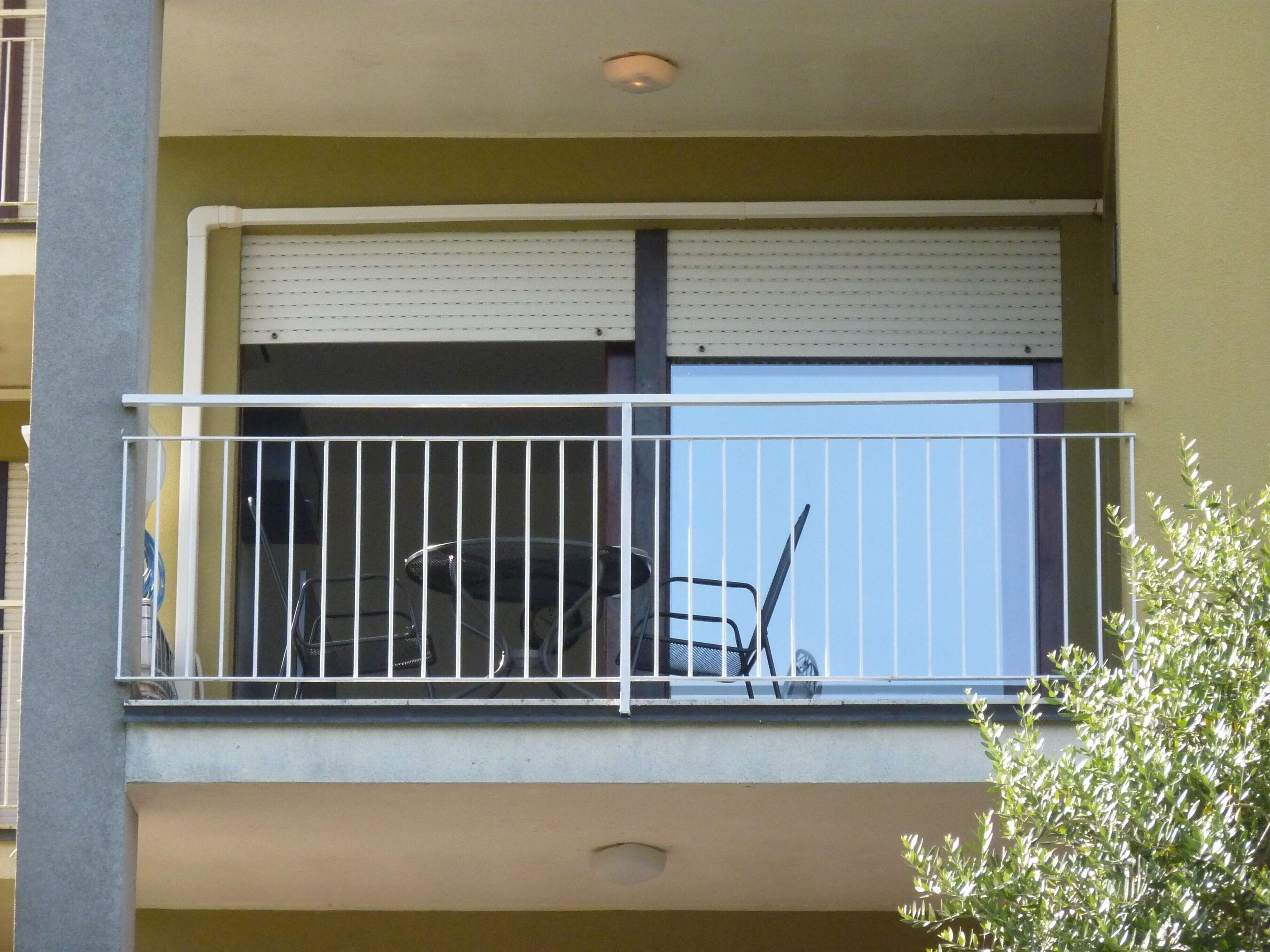 Un balcon avec une rambarde en métal, une fenêtre avec des stores et une petite plante.