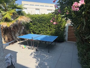 A ping pong table in an outdoor area with a tiled floor, surrounded by greenery and a building in the background. - Large family home with spacious, landscaped terraces, 100 meters from the beach (Mauguio)