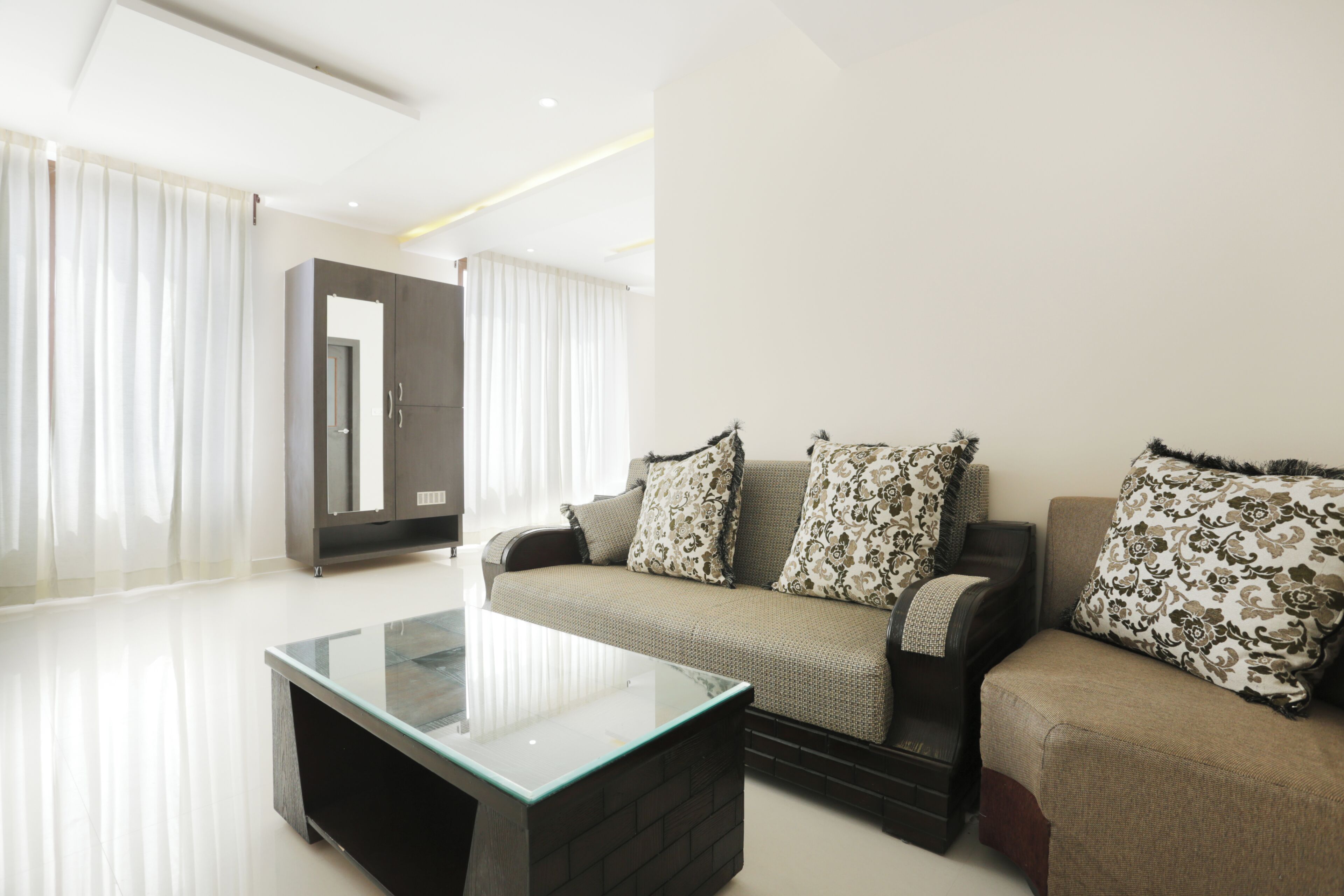 A modern living room with a glass coffee table, two sofas with patterned cushions, a wardrobe, and sheer curtains.