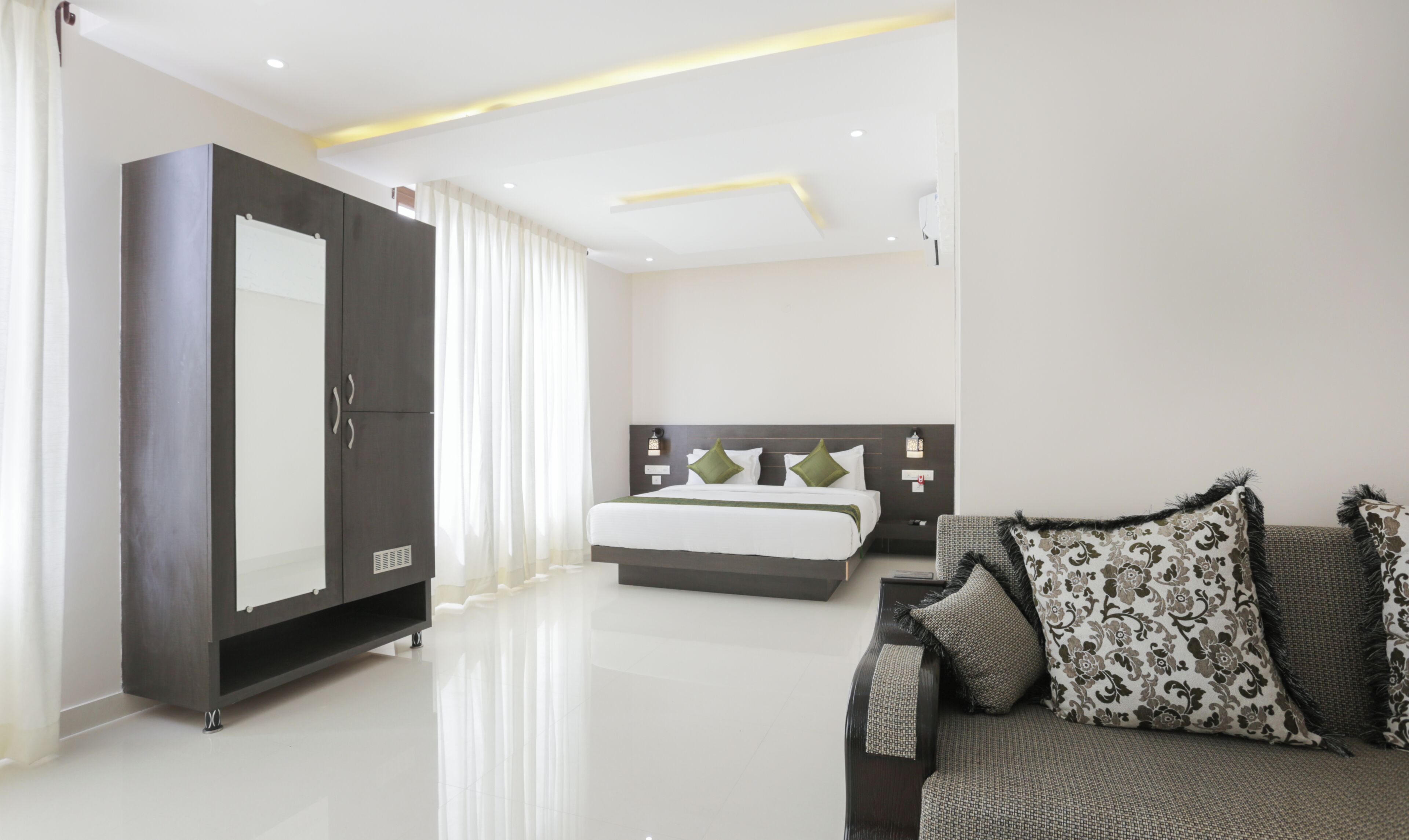 A modern hotel room with a large wardrobe, a bed with decorative pillows, a sofa with patterned cushions, and a clean, white floor.