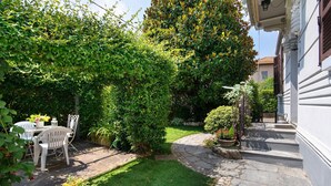 Un chemin de pierre mène à un bâtiment blanc doté d’un jardin comprenant une table et des chaises, une pelouse et diverses plantes.
