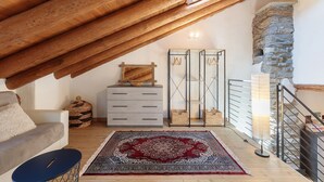 A loft bedroom with a sloped ceiling, a stone wall, a wooden ceiling beam, a grey dresser, a woven basket, a hanging rack, a red patterned rug, and a blue chair.