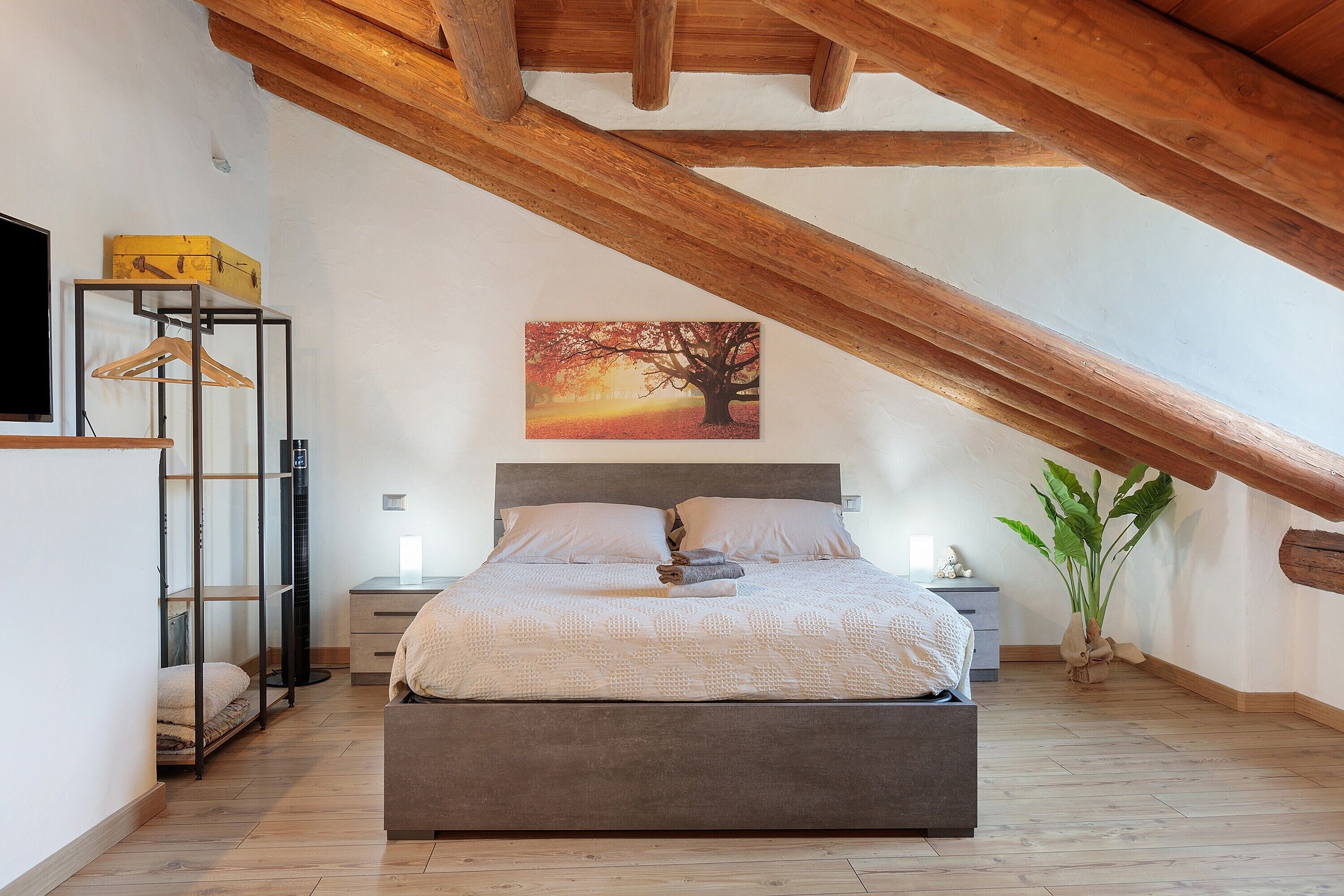 A bedroom with a bed, a nightstand, a bedside table, a hanging light, a painting, a plant, and a wooden ceiling beam.