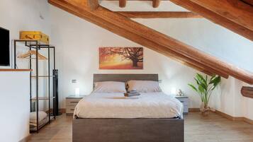 A bedroom with a bed, a nightstand, a bedside table, a hanging light, a painting, a plant, and a wooden ceiling beam.