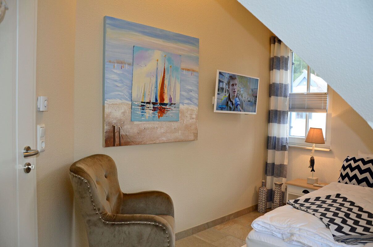 Ein Schlafzimmer mit einem Bett, einem Sessel, einem Bild von Segelbooten und einem Fenster mit Vorhängen.