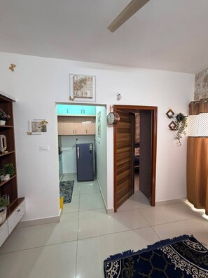Ein Zimmer mit Fliesenboden, einem blauen Teppich mit Muster, einer Holztür und einer Küchenzeile mit Schränken und einem Kühlschrank.
