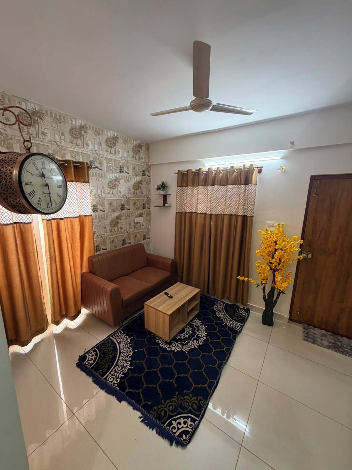 A living room with a sofa, a coffee table, a wall clock, curtains, a ceiling fan, and a patterned rug.