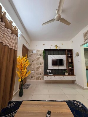 Ein Wohnzimmer mit Flachbildfernseher an der Wand, Deckenventilator, Holz-Couchtisch und einer Zierpflanze.