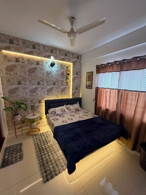 A bedroom with a bed, a ceiling fan, a wall-mounted light, a patterned wallpaper, a potted plant, and a window with curtains. - Home Away from Home – 1 BHK Apartment in Whitefield (Bengaluru)
