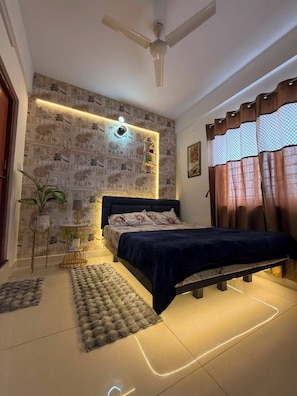 A bedroom with a bed, a ceiling fan, a wall-mounted light, a patterned wallpaper, and a window with curtains. - Home Away from Home – 1 BHK Apartment in Whitefield (Bengaluru)
