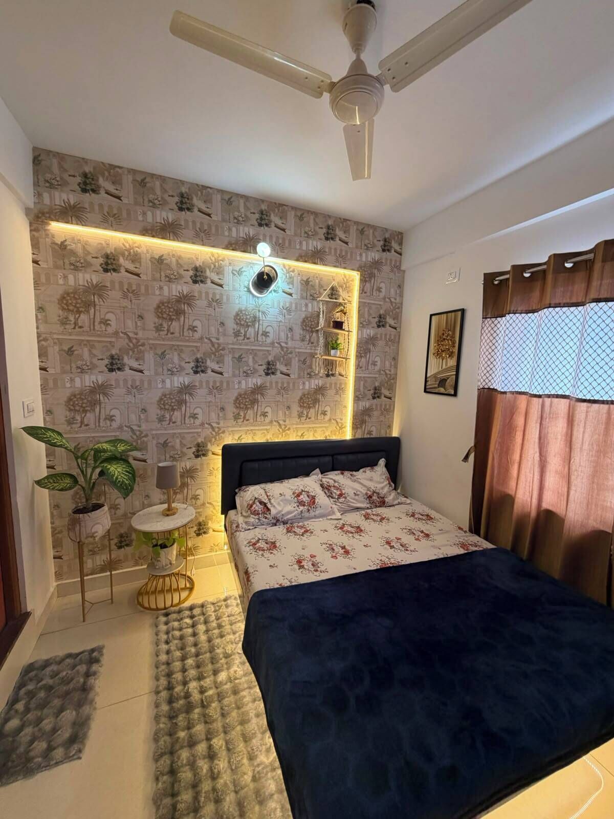 A bedroom with a bed, a ceiling fan, a wall-mounted shelf with plants, and a patterned wallpaper.