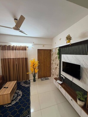 A modern living room with a flat-screen TV mounted on a wall, a ceiling fan, a wooden door, and a patterned rug. - Home Away from Home – 1 BHK Apartment in Whitefield (Bengaluru)