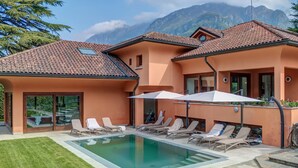 Un edificio a due piani con piscina, lettini e uno sfondo montuoso.