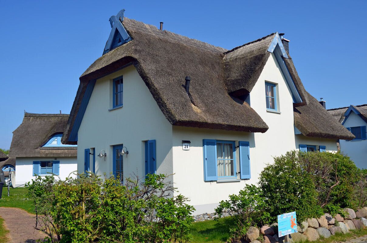 Une maison traditionnelle à toit de chaume, aux murs blancs et aux volets bleus, située au numéro 29 et entourée de verdure.