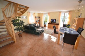 Ein Wohnzimmer mit Treppe, einer Couch, einem Esstisch mit Stühlen, einem Fernseher und einem Bücherregal.