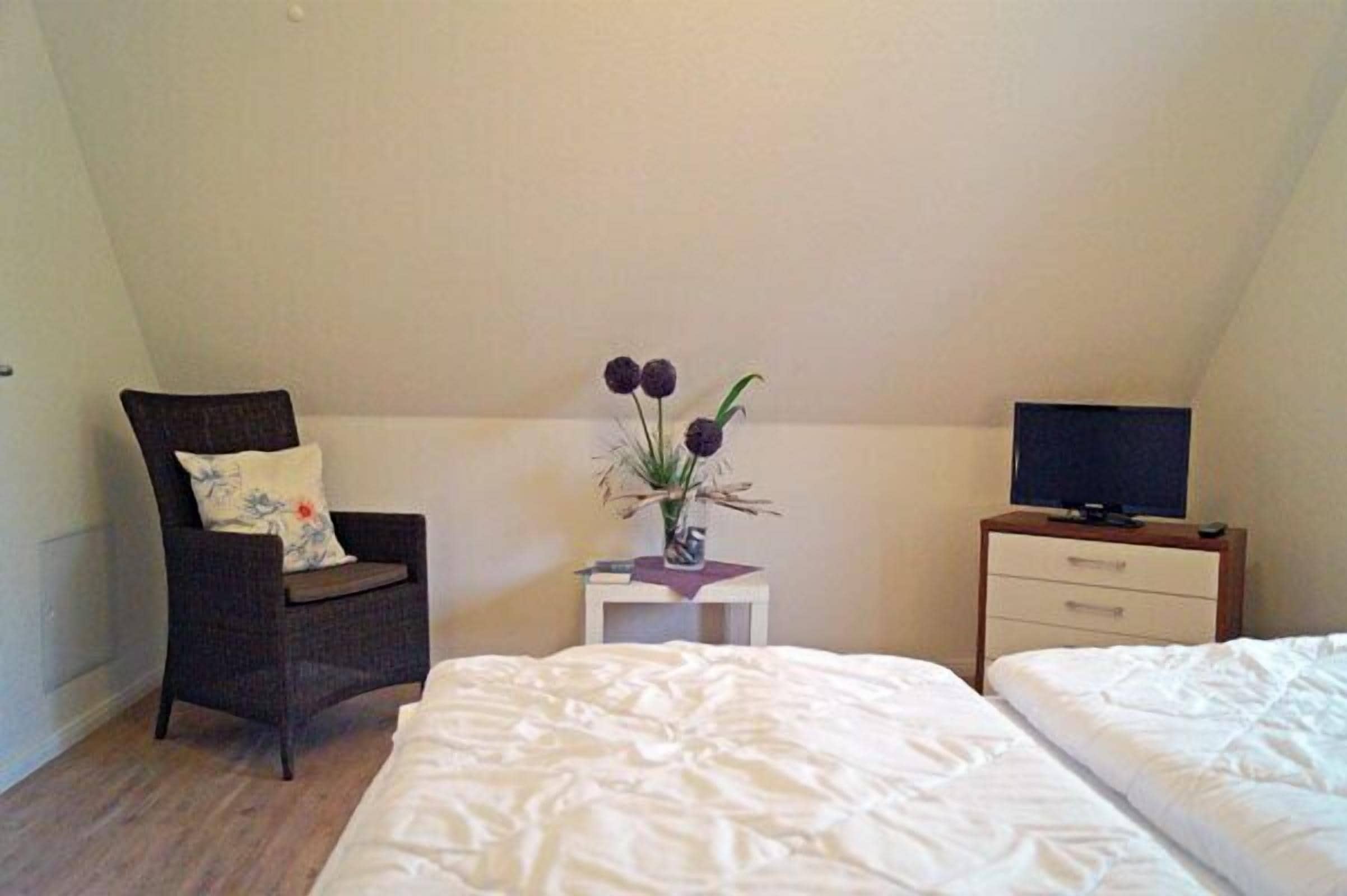 A bedroom with a bed, a chair, a small table with a vase, and a TV on a stand.
