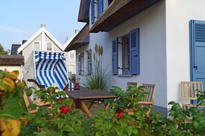Een gebouw in traditionele Europese stijl, met rieten dak, blauwe luiken en een houten tafel met stoelen op de voorgrond.