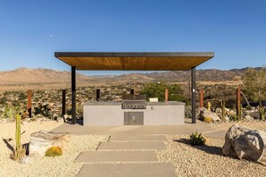 An outdoor barbecue area with a grill, surrounded by desert plants and a view of a distant town and mountains. - Rattlesnake Ranch - Hollywood Retreat w Pool&Views (Yucca Valley)