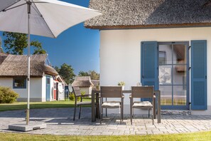Eine Terrasse mit Tisch und Stühlen, ein Gebäude mit einem Reetdach und ein weißer Sonnenschirm.