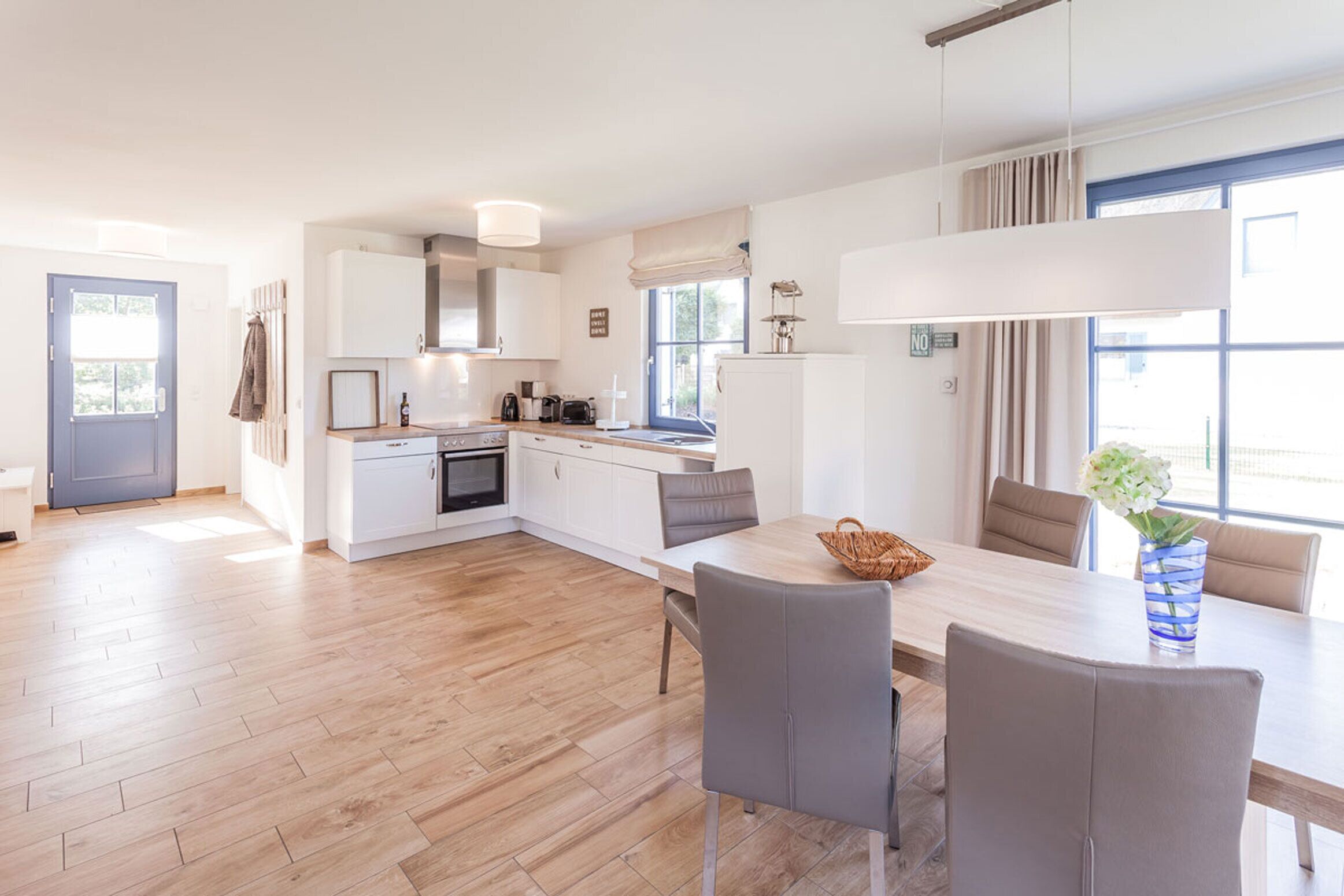 A modern kitchen with white cabinetry, a dining area with a table and chairs, and a view of the outdoors through large windows.
