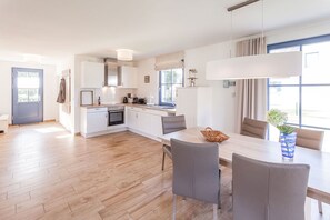Eine moderne Küche mit weißen Schränken, einem Essbereich mit Tisch und Stühlen und Blick ins Freie durch große Fenster.