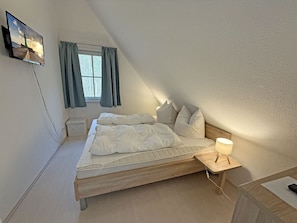 A bedroom with a bed, a bedside table with a lamp, a window with curtains, and a TV mounted on the wall. - Vacation home with a fireplace, about 800 meters from the sandy beach, with a washing machine and Wi-Fi (Glowe)