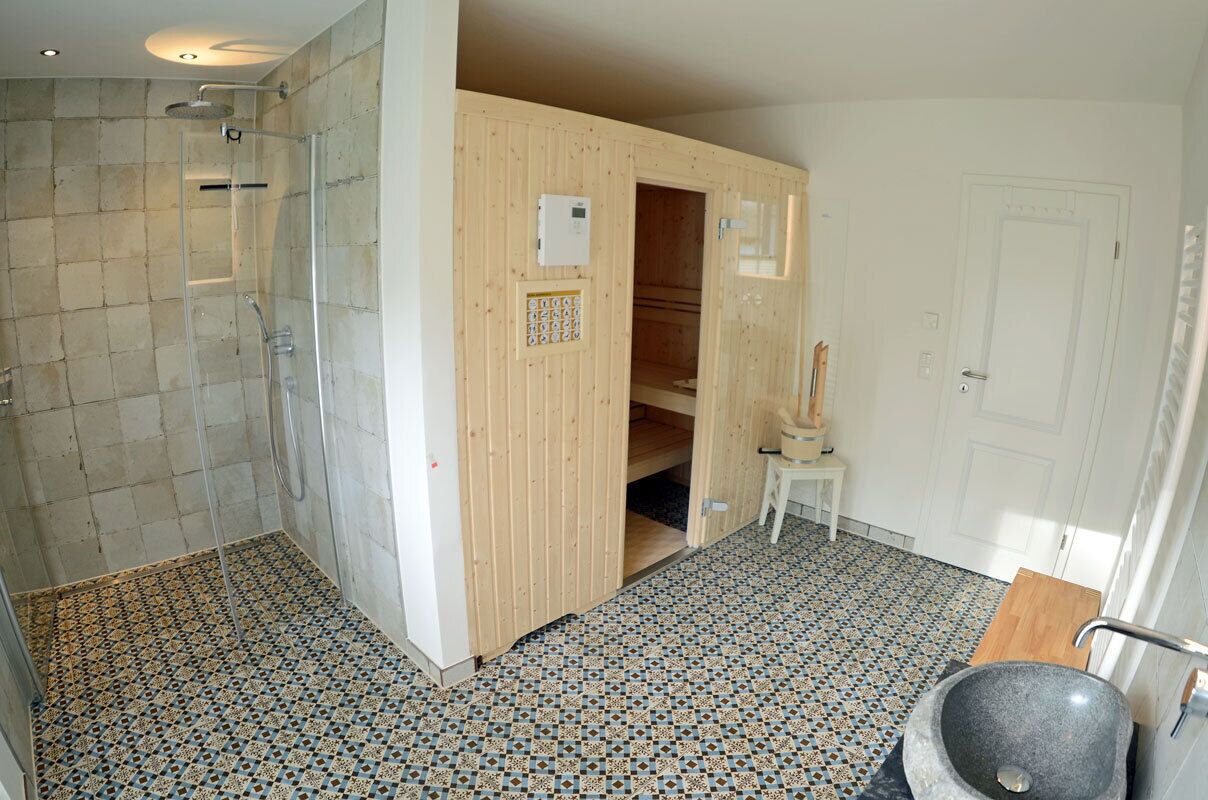 A bathroom with a shower, a wooden sauna, a small white stool, and a stone sink.