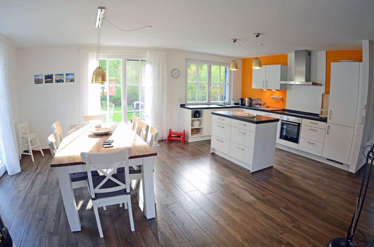 A modern kitchen with white cabinets, a wooden dining table, and a view of the outdoors through large windows.
