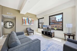 Ein modernes Wohnzimmer mit einem grauen Sofa, einem Fernseher, einem Gemälde und Blick auf die Stadt durch große Fenster.