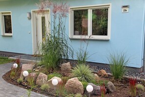Ein Wohngebäude mit hellblauer Fassade, der Hausnummer '23a' an der Wand und einem angelegten Garten mit Steinen und Pflanzen.