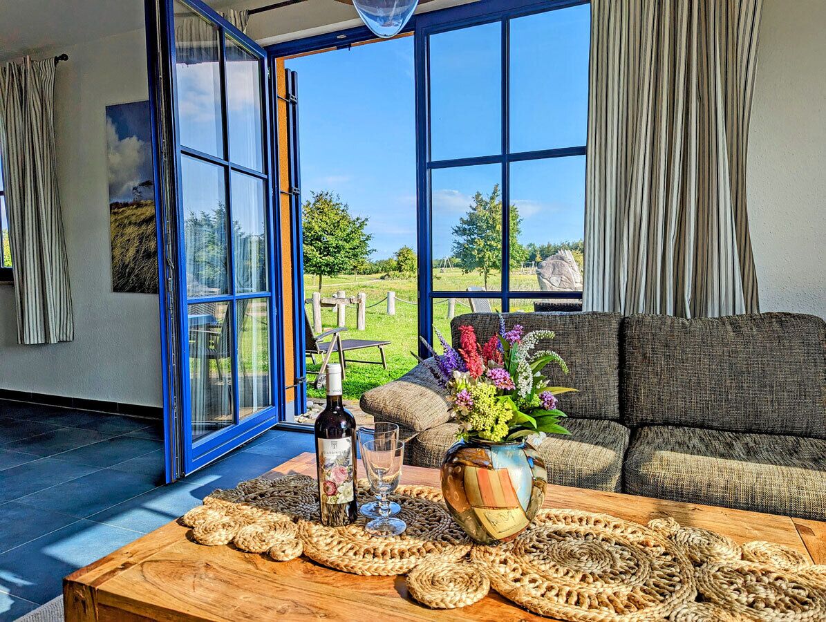 A room with a large window, a sofa, a wooden coffee table with woven placemats, a bottle of wine, and a vase with flowers.
