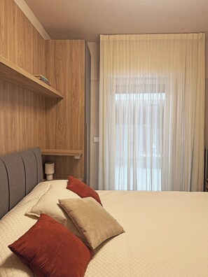 Ein Hotelzimmer mit Bett, Kissen, einem Nachttisch mit Lampe, einer Holztrennwand und einem Fenster mit Vorhängen.