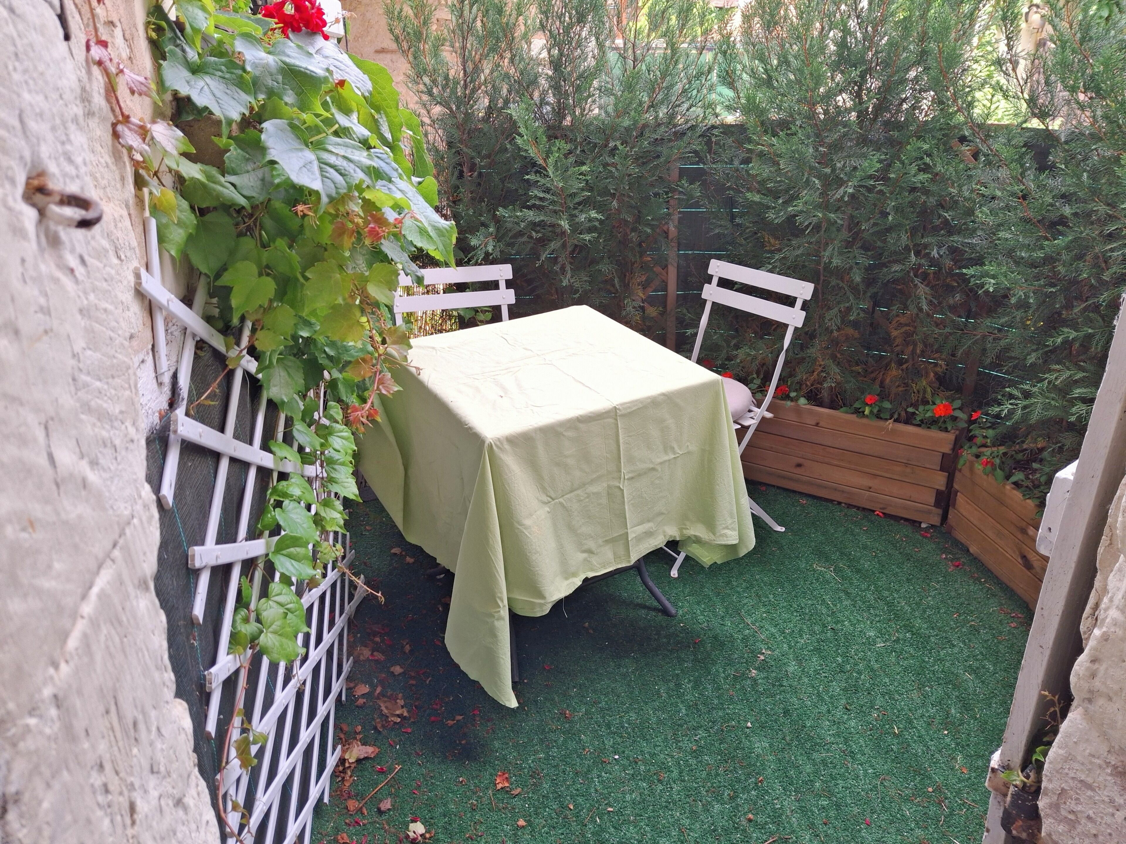 Un pequeño espacio al aire libre con una mesa y sillas, rodeado de follaje y una jardinera de madera con flores rojas.