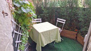 A small outdoor seating area with a table and chairs, surrounded by greenery and a wooden planter with red flowers. - TOWNHOUSE, 2 minutes from the river, with shops and a small courtyard filled with flowers (Barrou)