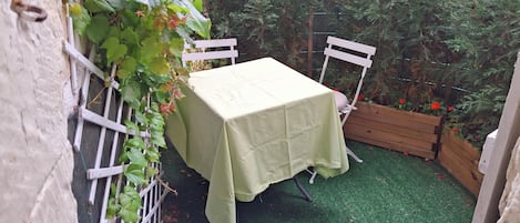 Un pequeño espacio al aire libre con una mesa y sillas, rodeado de follaje y una jardinera de madera con flores rojas.