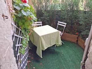 Ein kleiner Sitzbereich im Freien mit Tisch und Stühlen, umgeben von Pflanzen und einem Holzbeet mit roten Blumen.