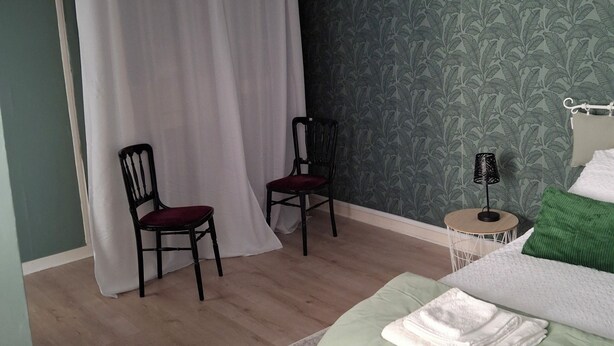 A bedroom with a bed, two chairs, a small table with a lamp, and a curtain. - TOWNHOUSE, 2 minutes from the river, with shops and a small courtyard filled with flowers (Barrou)