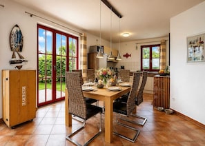 A dining area with a wooden table set for four, surrounded by wicker chairs, a kitchen in the background, and a view of the outdoors through large windows. - Vacation home with a fireplace, about 250 meters from the sandy beach, Wi-Fi, pets welcome (Glowe)