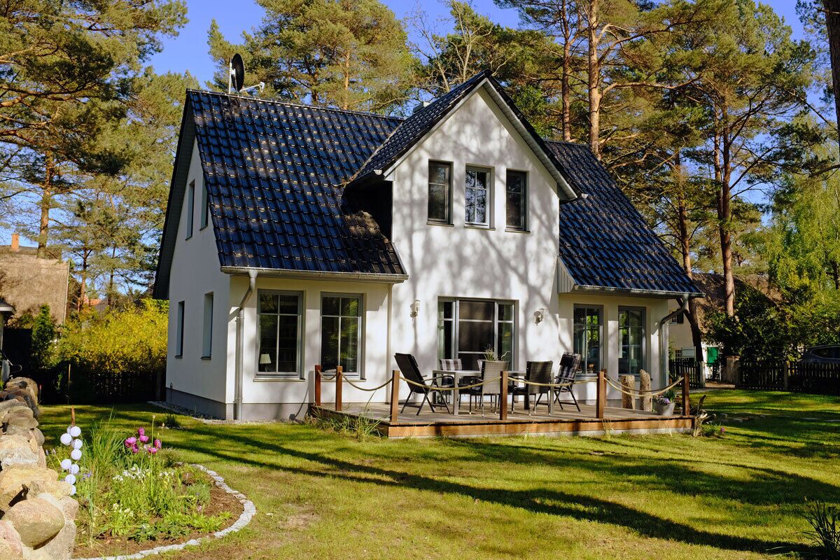 A two-story house with a sloped roof, a covered porch with chairs, and a garden with flowers and a stone wall.
